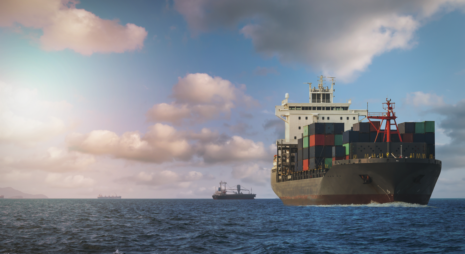 Container ship sailing on open water under cloudy sky, with two other vessels visible in the distance. The ship is loaded with multicolored shipping containers, showing typical maritime commercial operations.